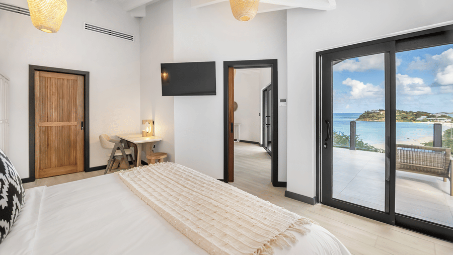 A bedroom with a king size bed, lamps, a door, a television, and glass doors leading to a terrace with an ocean view at Tamarind Hills Resort and Villas, Antigua.
