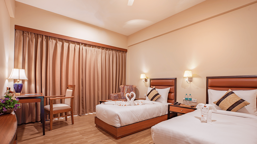 A spacious bedroom at The Lalita Grand Mathura - Vrindavan featuring twin beds, decorative towel art, wooden furniture, warm lighting, and long curtains.