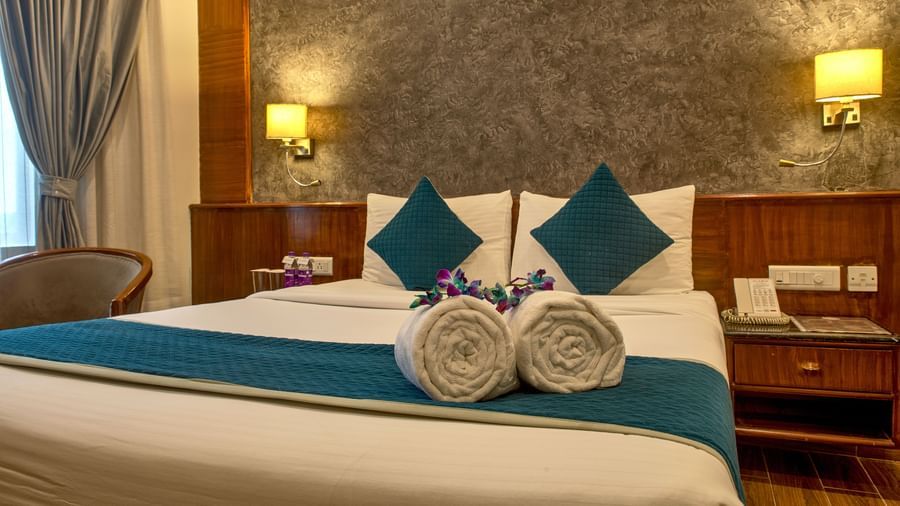 Comfortable bed with teal cushions and neatly folded towels arranged in a flower shape at Ilara Hotels & Spa, OMR, Chennai
