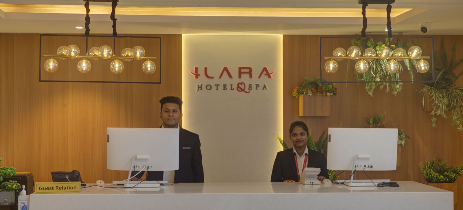 Hotel reception area with two front desk staff members standing behind a modern white counter beneath elegant hanging lights at Ilara Hotels & Spa.