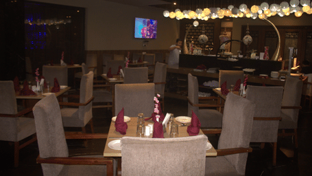 Indoor restaurant with grey upholstered dining chairs, square wooden tables, maroon napkins, and pendant lighting above the service counter at Pipul Padmaja Premium Hotel & Convention.