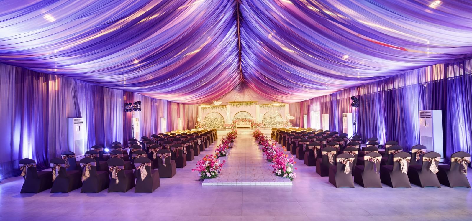 Elegant banquet hall with decorated roof in purple colour and rows of black covered chairs with flower centrepieces along the isle at Grande Bay Resort & Spa.