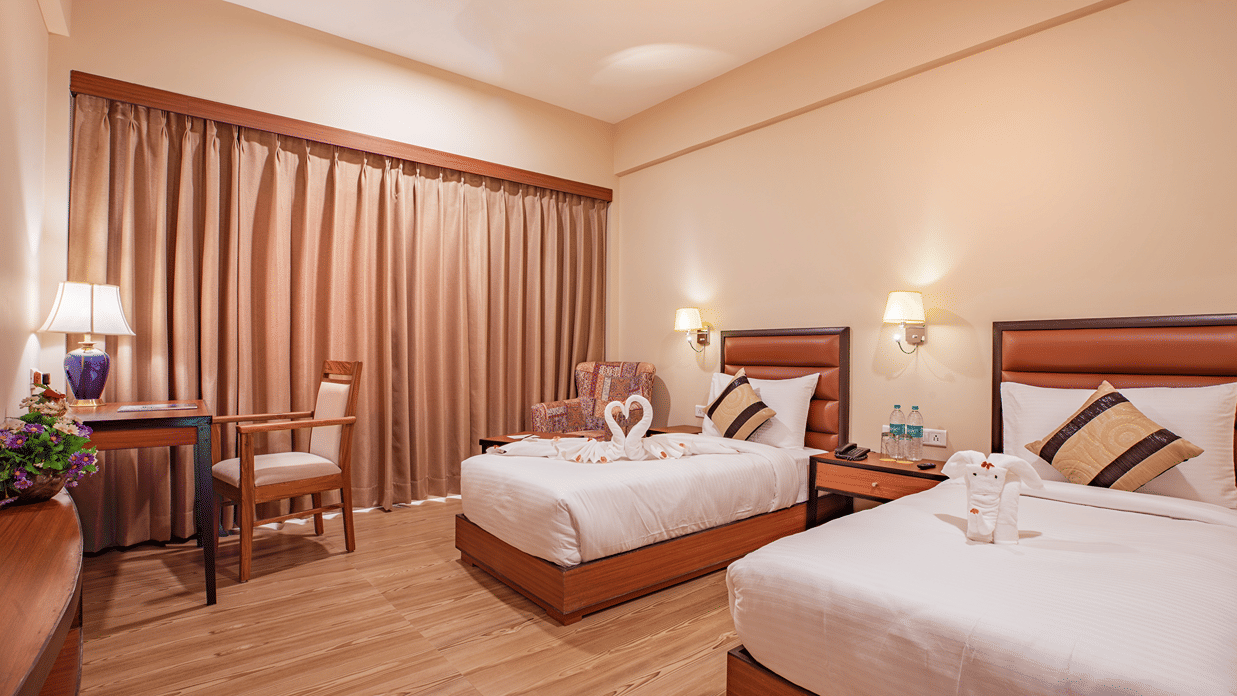 A spacious bedroom at The Lalita Grand Mathura - Vrindavan featuring twin beds, decorative towel art, wooden furniture, warm lighting, and long curtains.