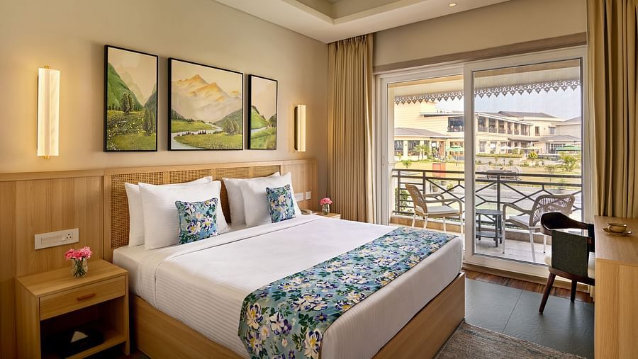 A hotel bedroom with a king bed dressed in white and a blue floral runner. Three landscape paintings hang above the wooden headboard. A nightstand with flowers sits beside the bed. Sliding glass doors with light curtains open to a balcony - Voyage The Soli