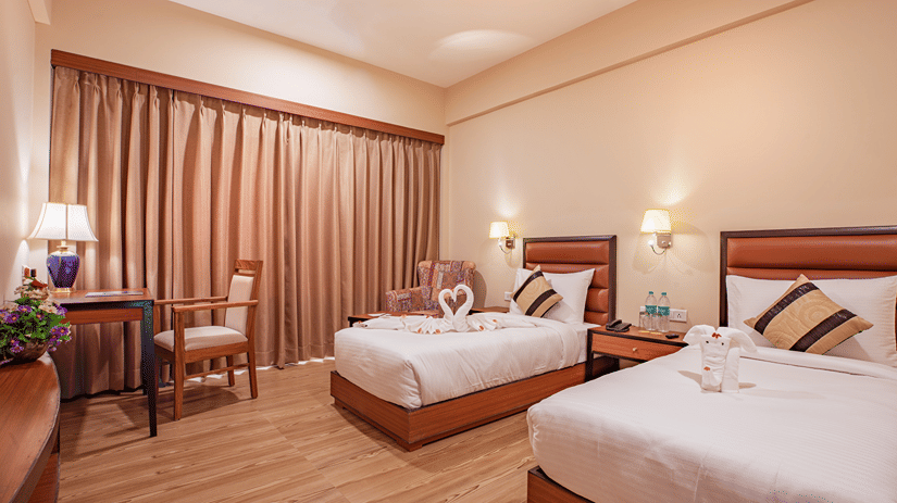A spacious bedroom at The Lalita Grand Mathura - Vrindavan featuring twin beds, decorative towel art, wooden furniture, warm lighting, and long curtains.