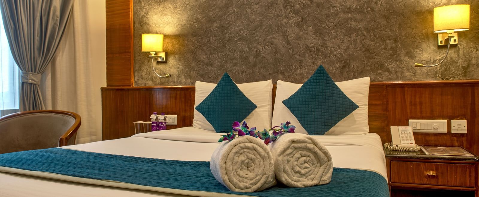 Comfortable bed with teal cushions and neatly folded towels arranged in a flower shape at Ilara Hotels & Spa, OMR, Chennai
