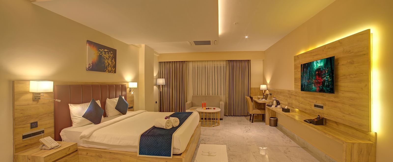 A warmly lit bedroom of the Junior Suite at Ilara Hotels & Spa, Tada featuring a double bed and wall mounted television in front of it.
