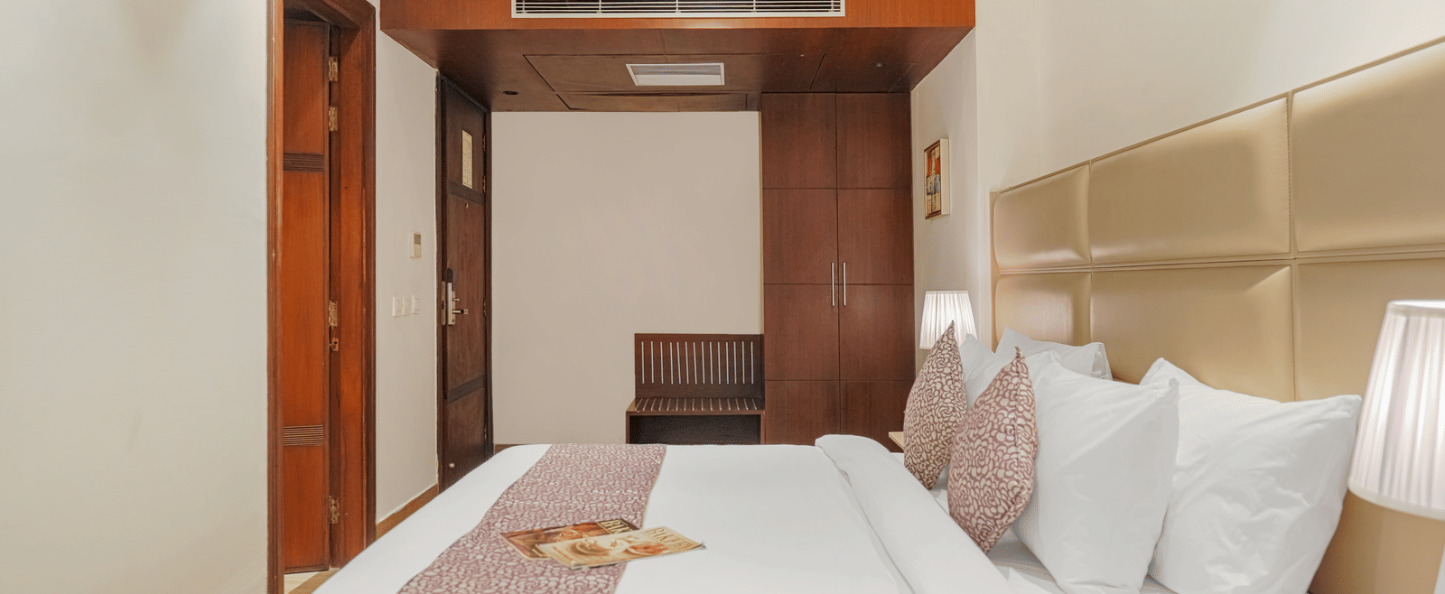 A bright hotel bedroom showing a large bed with a patterned runner and an integrated, open wooden wardrobe at BluSalzz Select City Centre, Amritsar.