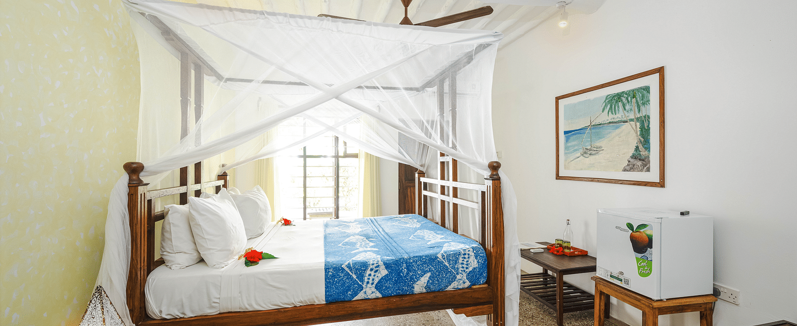 Interior view of a bedroom with a wooden four-poster bed with a mosquito net, a patterned bedspread, a framed picture on the wall, a small refrigerator, and a wooden table at shooting star Boutique Hotel.