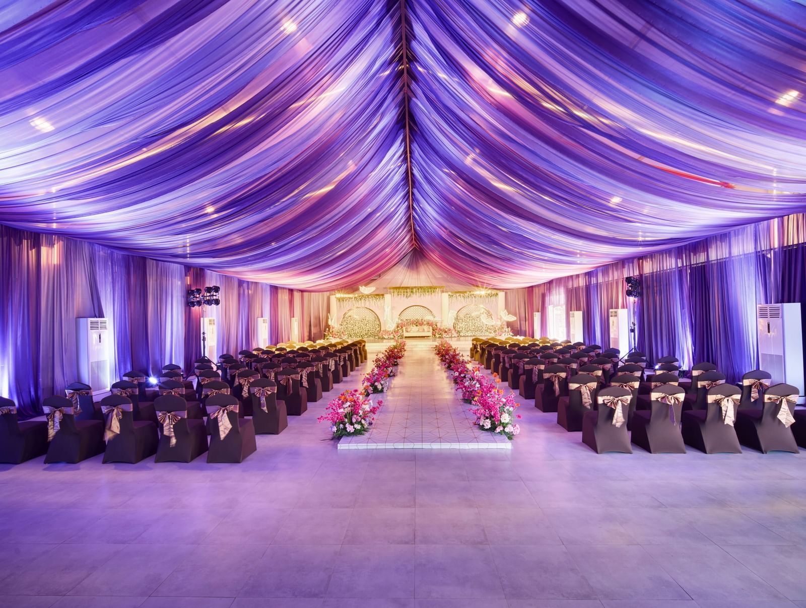 Elegant banquet hall with decorated roof in purple colour and rows of black covered chairs with flower centrepieces along the isle at Grande Bay Resort & Spa.