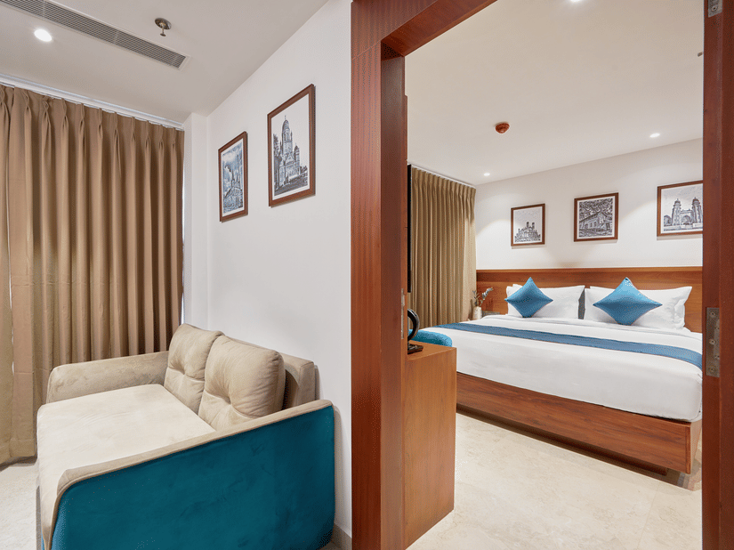View of a suite at QWhite Crescent Luxury Hotel by Dimora Kochi with a connecting living area, teal sofa, king bed, and warm ambient lighting.