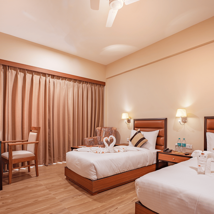 A spacious bedroom at The Lalita Grand Mathura - Vrindavan featuring twin beds, decorative towel art, wooden furniture, warm lighting, and long curtains.