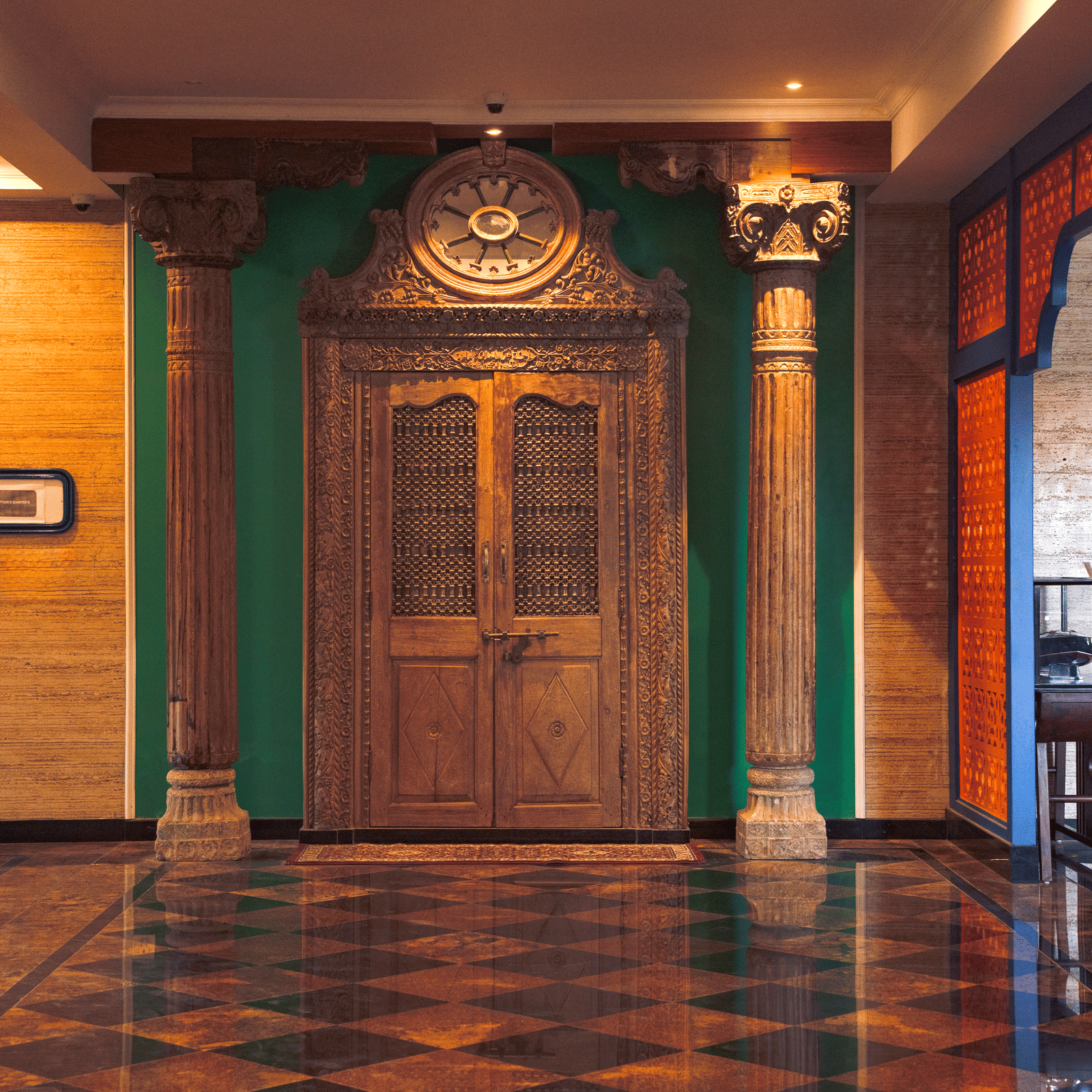 A grand corner hallway with polished flooring, carved wooden double doors, tall columns, wall decor and warm lighting creating a traditional welcome area at MAYFAIR Bay Resort, Paradeep.