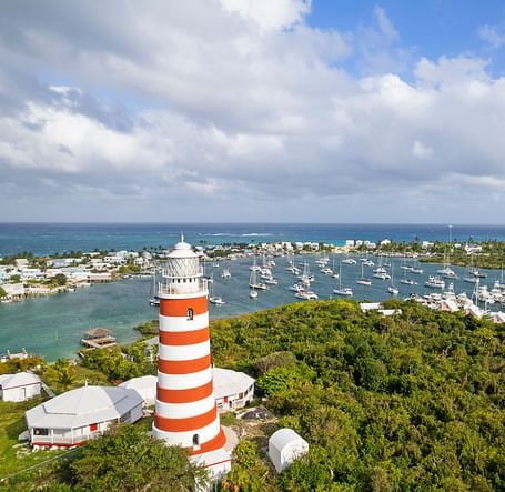 An aerial view of Hope Town, showing a red-and-white striped lighthouse, marina, and buildings surrounded by greenery and blue waters | Abaco Inn