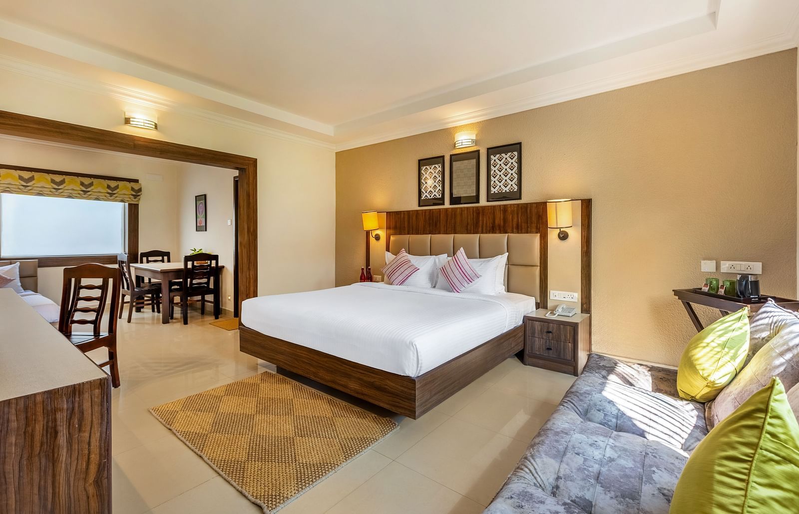 A hotel room with a queen bed, a sofa, and a doorway leading to a dining area with a table and chairs - Mango Hill Resort, Coimbatore