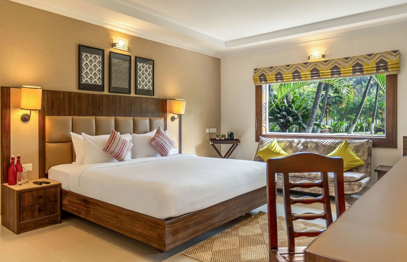 A hotel room with a bed, a desk, a sofa, and a large window overlooking a garden with palm tree - Mango Hill Resort, Coimbatore