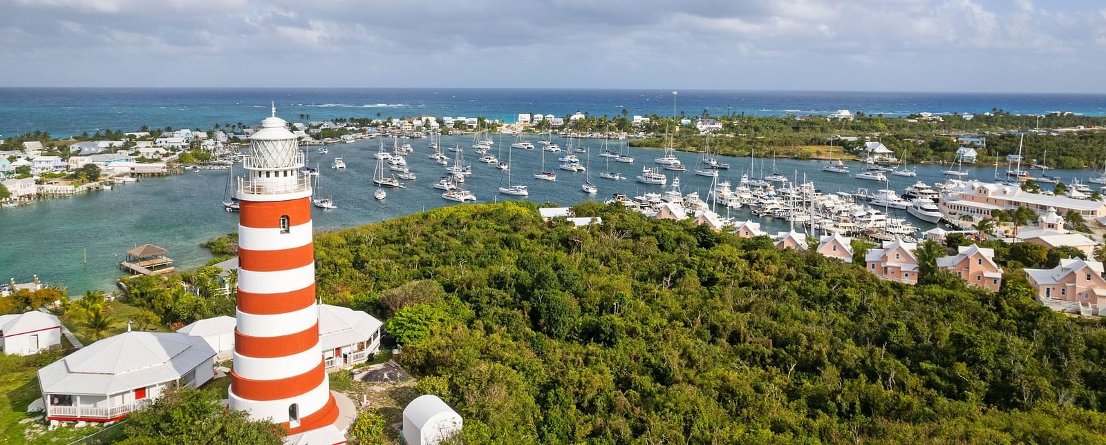 An aerial view of Hope Town, showing a red-and-white striped lighthouse, marina, and buildings surrounded by greenery and blue waters | Abaco Inn
