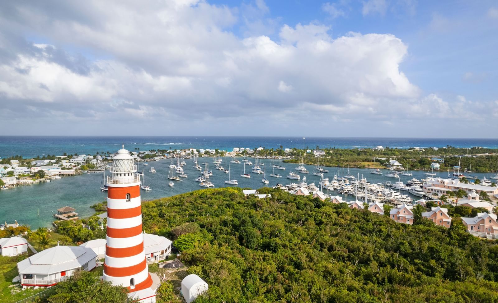 An aerial view of Hope Town, showing a red-and-white striped lighthouse, marina, and buildings surrounded by greenery and blue waters | Abaco Inn