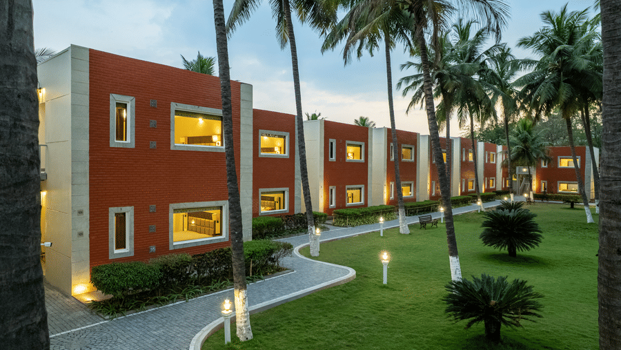 A building with red brick accents and white trim, surrounded by palm trees and a paved pathway - Mango Hill Resort, Coimbatore