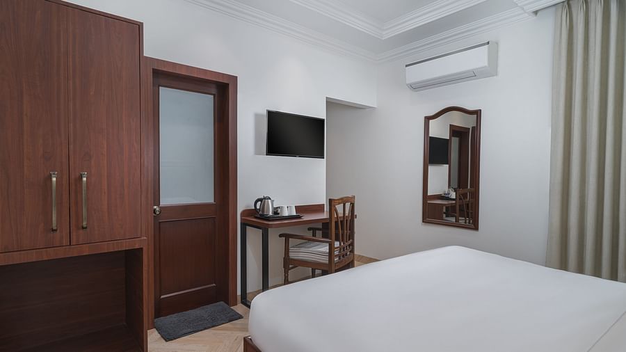 A room with a spacious bed, a wooden wardrobe, a wall-mounted television, and a wooden table directly below it | Hotel Ganga Maiya