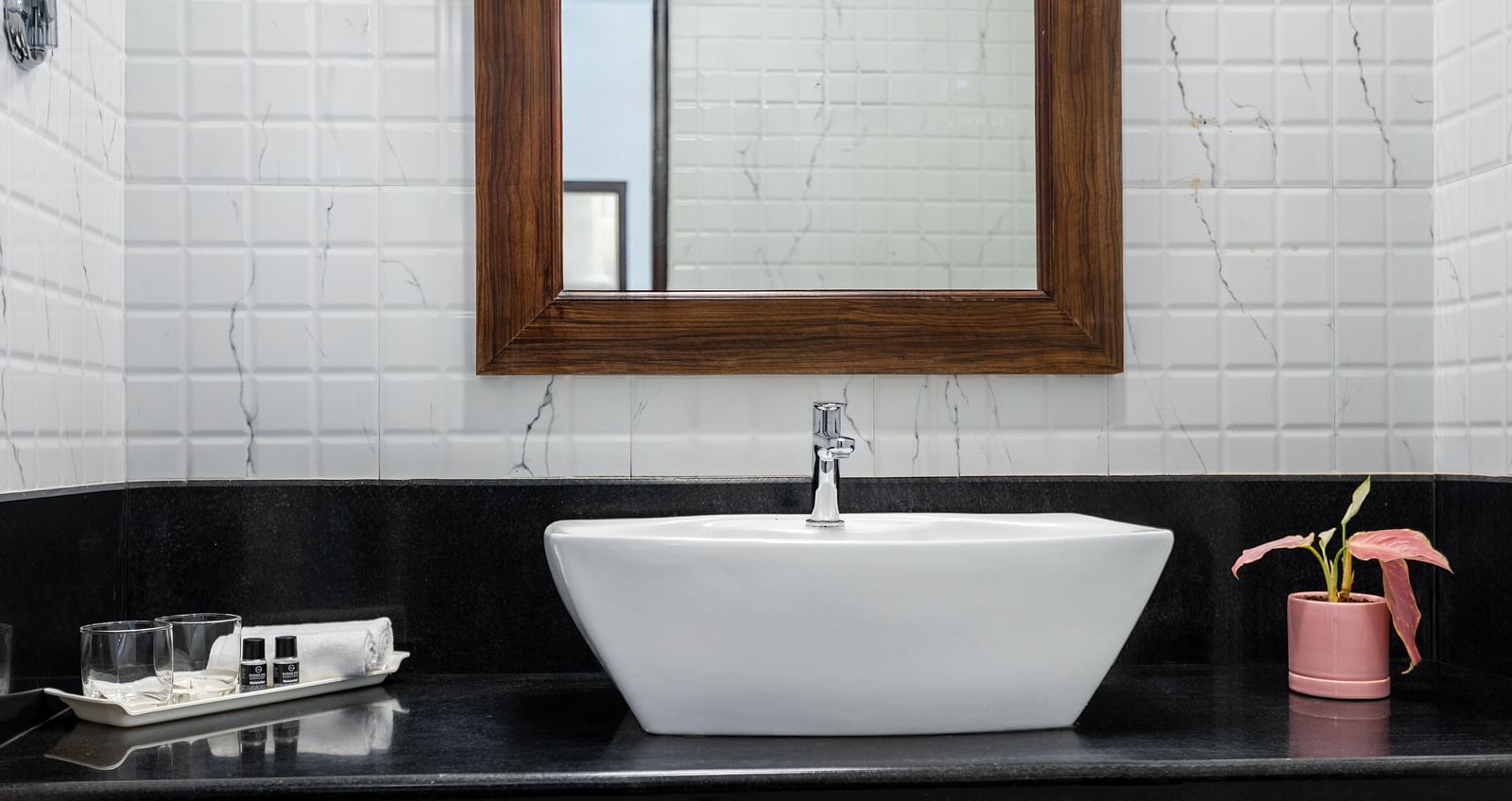 A bathroom with a white rectangular sink, a wooden framed mirror, white tiled walls, and a black countertop - Mango Hill Resort, Coimbatore