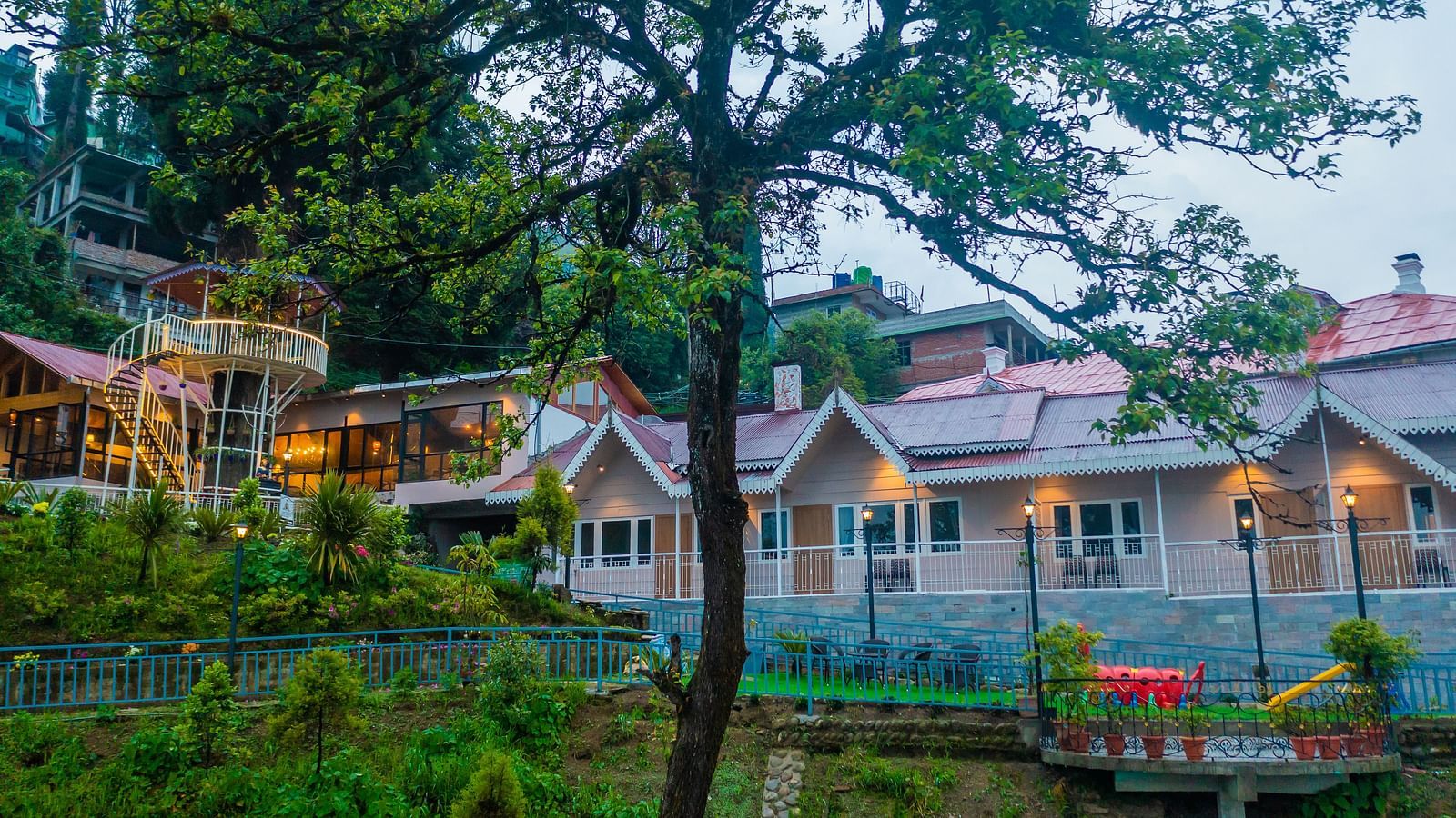  Hillside cottages with glowing lights at dusk, nestled among verdant foliage, showcasing a blend of natural tranquillity and cosy accommodation- Arcadia Heritage Resort, Darjeeling