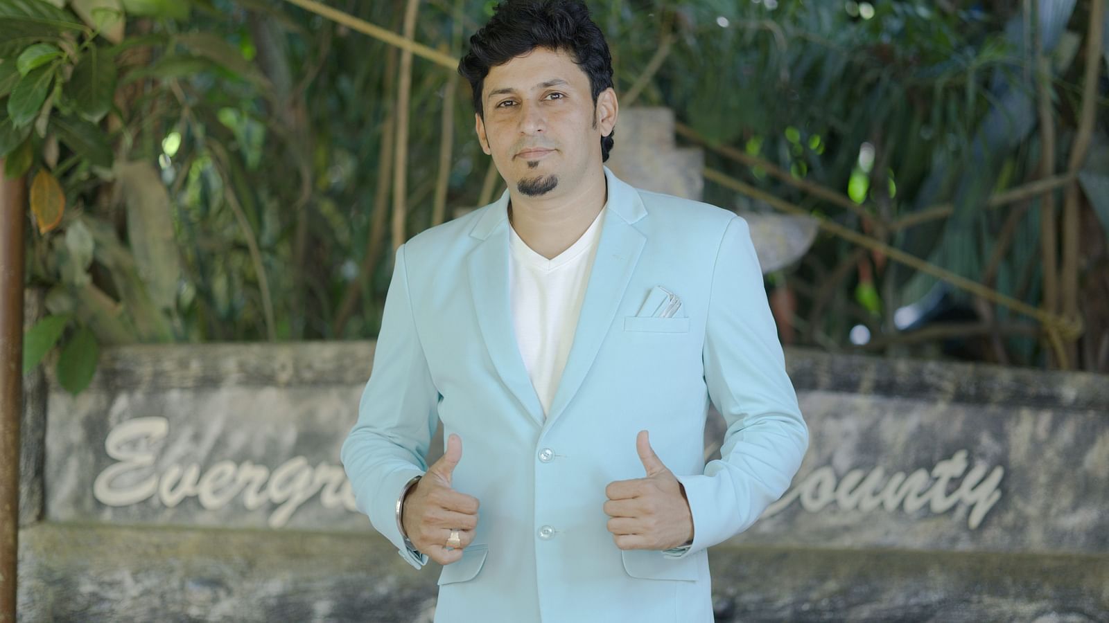 Man in a light blue suit posing confidently in front of a rustic wooden sign that reads Evergreen County, surrounded by lush greenery.