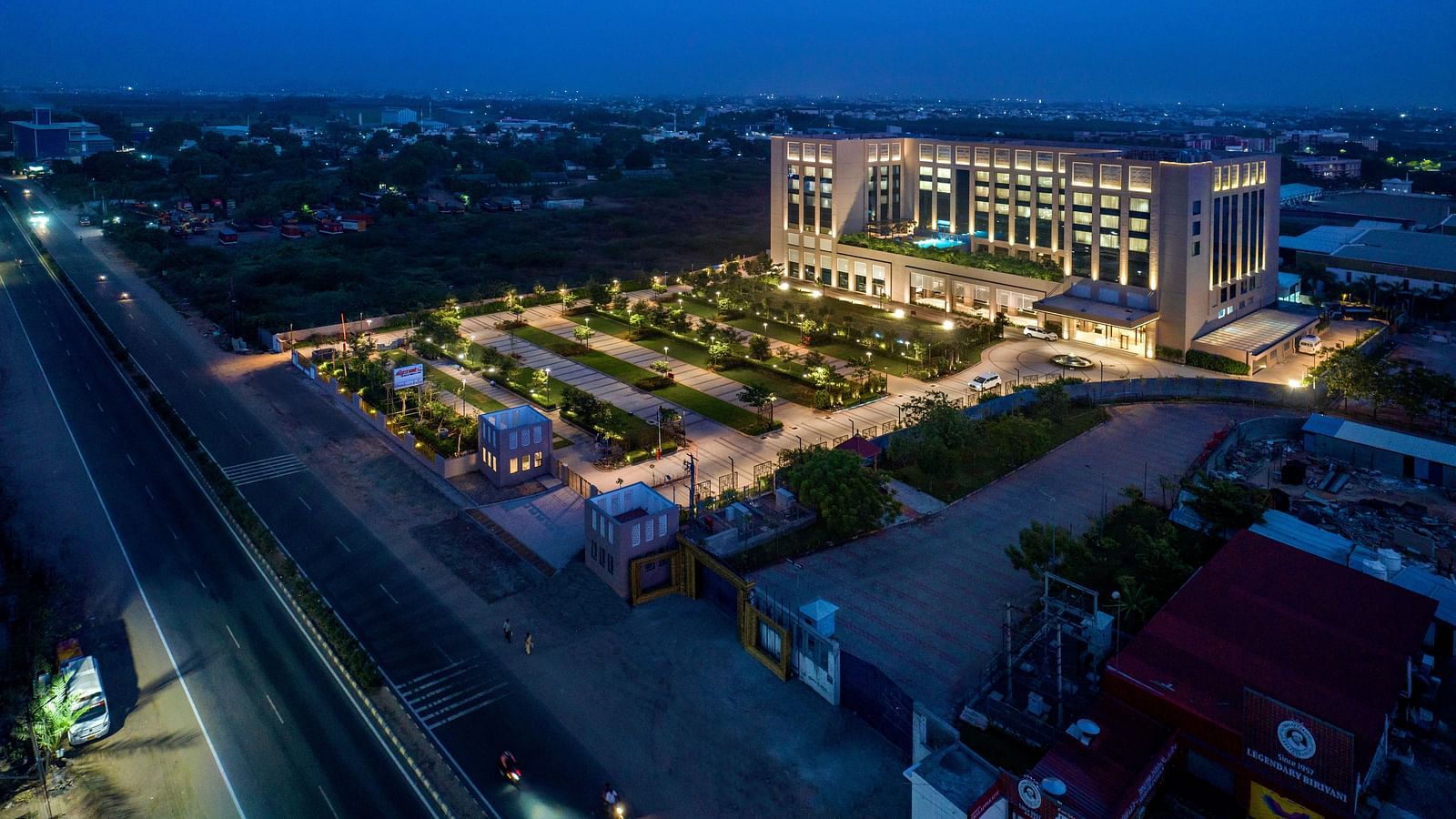An aerial view at dusk shows a large hotel building with illuminated grounds and a road - Grand Madurai by GRT Hotels