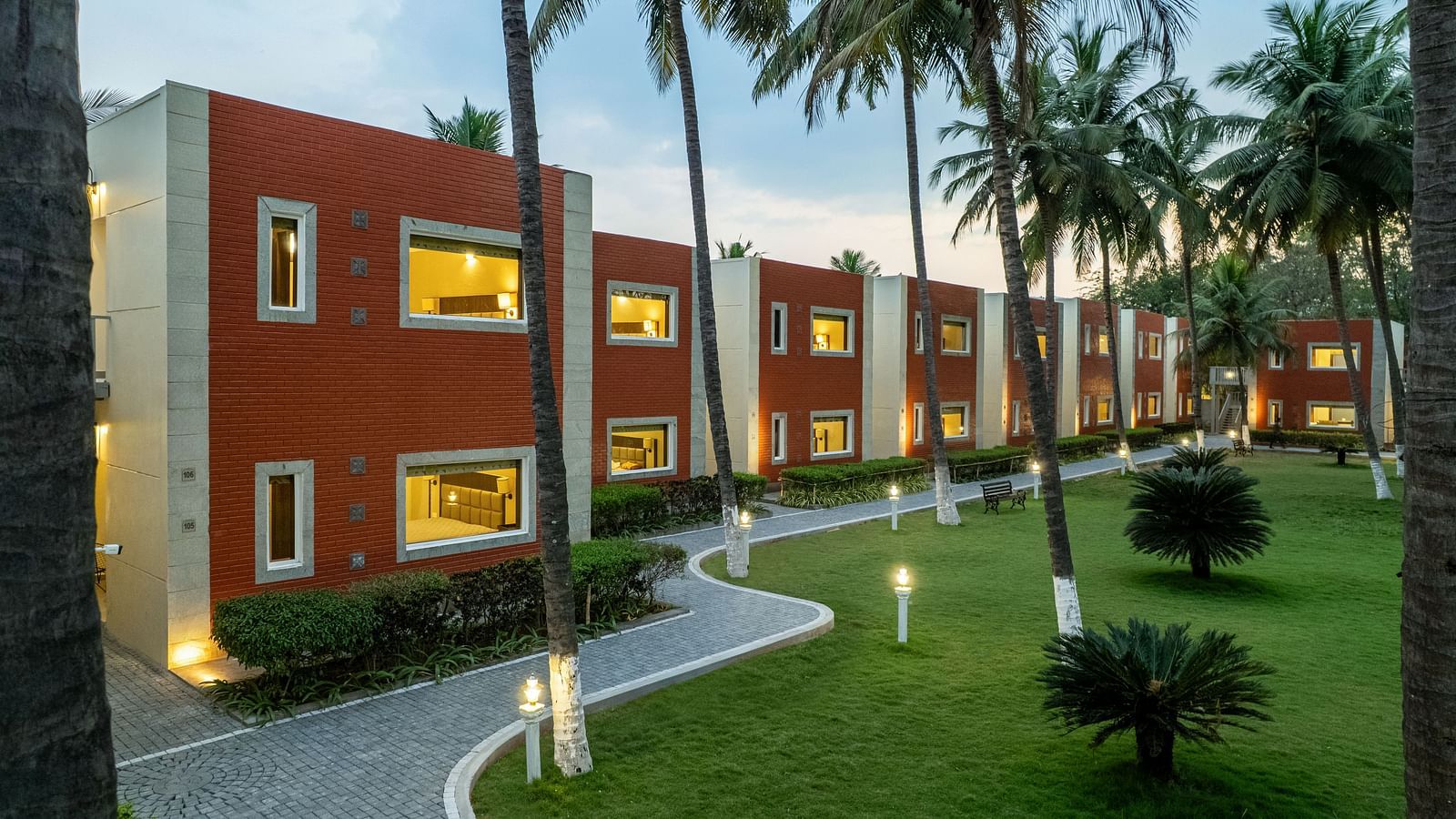 A building with red brick accents and white trim, surrounded by palm trees and a paved pathway - Mango Hill Resort, Coimbatore