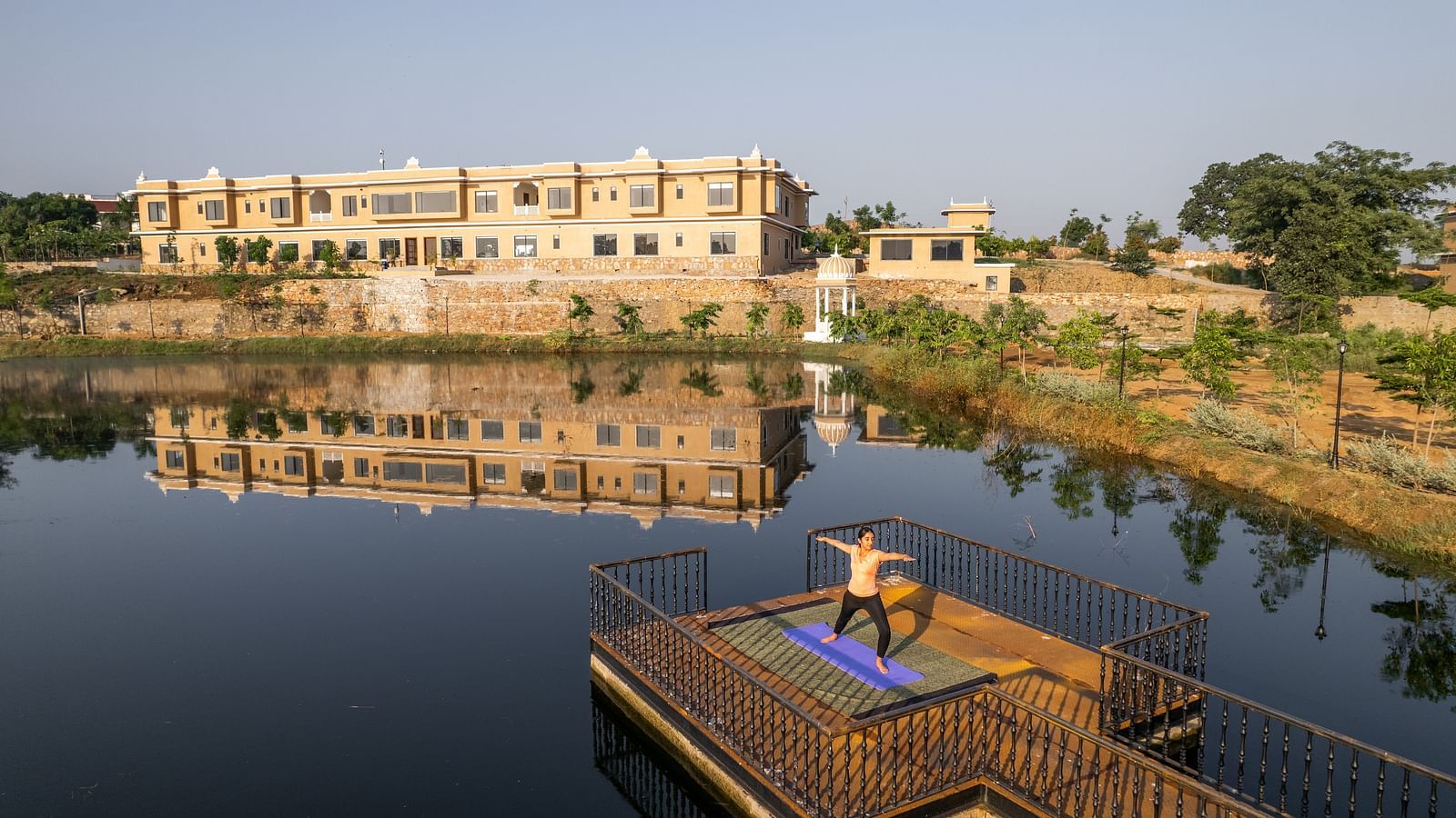 An image of a woman performing a yoga asan on a platform surrounded by water with our property facade in the back - Rajbagh Wellness, Udaipur