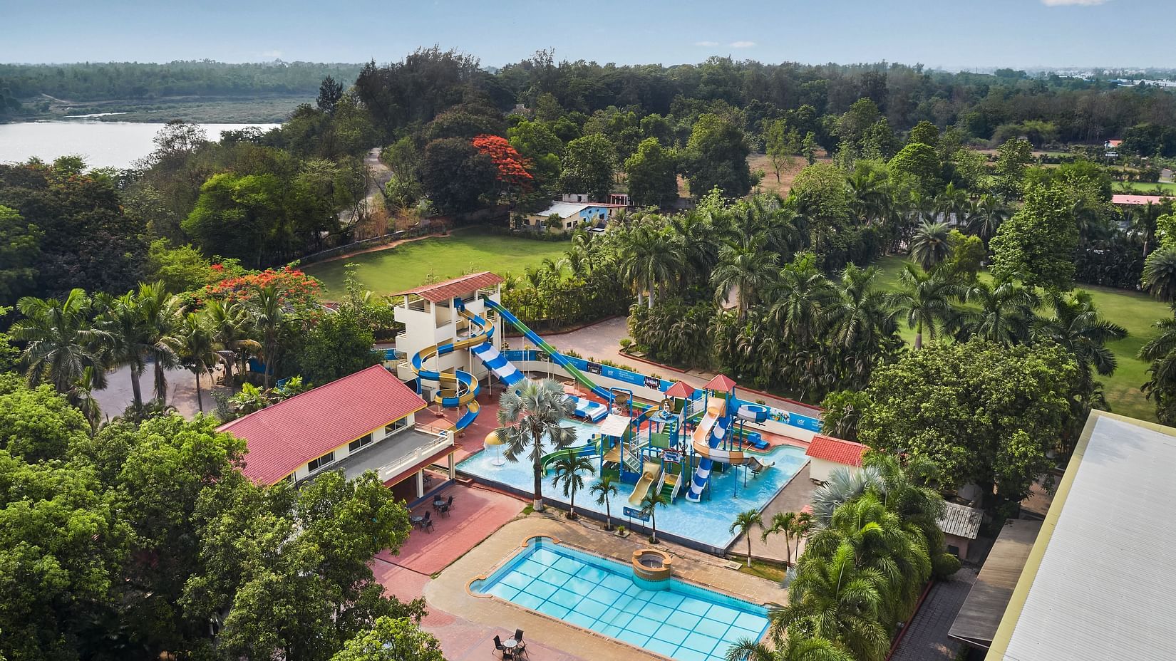 DGV Resort by Treat, Silvassa with a colourful waterpark featuring slides, splash areas, a swimming pool, and red-roofed buildings near a riverside