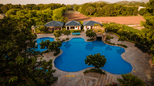 A swimming pool with curved edges surrounded by greenery pathways and villa-style buildings set within a wooded area at The Baagh Ananta Elite, Ranthambore.