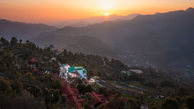 A landscape view at sunset or sunrise, showing hills and valleys with a few buildings illuminated in the fading light