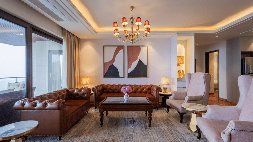 Elegant living room with a leather sofa, plush chairs, a vibrant chandelier, and large windows, presenting a sophisticated gathering area in The Manor Sports & Wellness Hotel in Shimla.