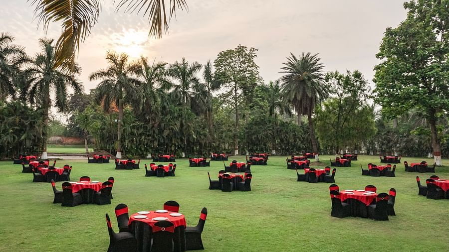 Outdoor lawn setup with round tables covered in red and black linen, surrounded by palm trees and greenery under a cloudy sky at DGV Resort by Treat, Silvassa