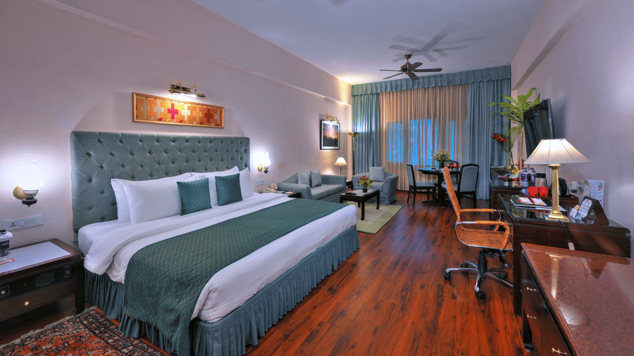 A room with a large bed, spreads, pillows, a fan, and windows at The Clarks Varanasi.