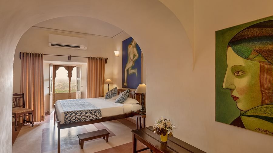 Hotel room with a bed, bedside tables with lamps, a bench, artwork, and a balcony with ornate railings at Tijara Fort-Palace, Alwar.