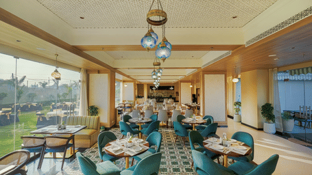 The well-lit Urban Rasoi at Urban Suites by BluSalzz, Jaipur, featuring upholstered chairs, wooden flooring, and large windows with an outdoor view.