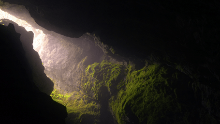 Sunlight streams through the entrance of a moss-covered cave, illuminating rugged rock formations and creating a mystical underground landscape.