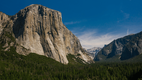 A panoramic view of a massive rock formation with a clear blue sky and forested valley below.