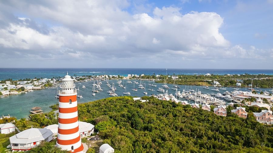 An aerial view of Hope Town, showing a red-and-white striped lighthouse, marina, and buildings surrounded by greenery and blue waters | Abaco Inn