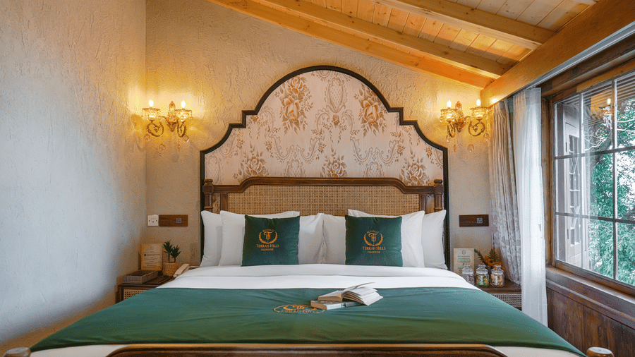 The claasic room at BluSalzz Terrah Hills Resort, Dalhousie, with a double bed featuring a bedspread, books, and pillows, wall mounted light fixtures, set against a backdrop of unique wall architecture, and a window view of lush greenery.