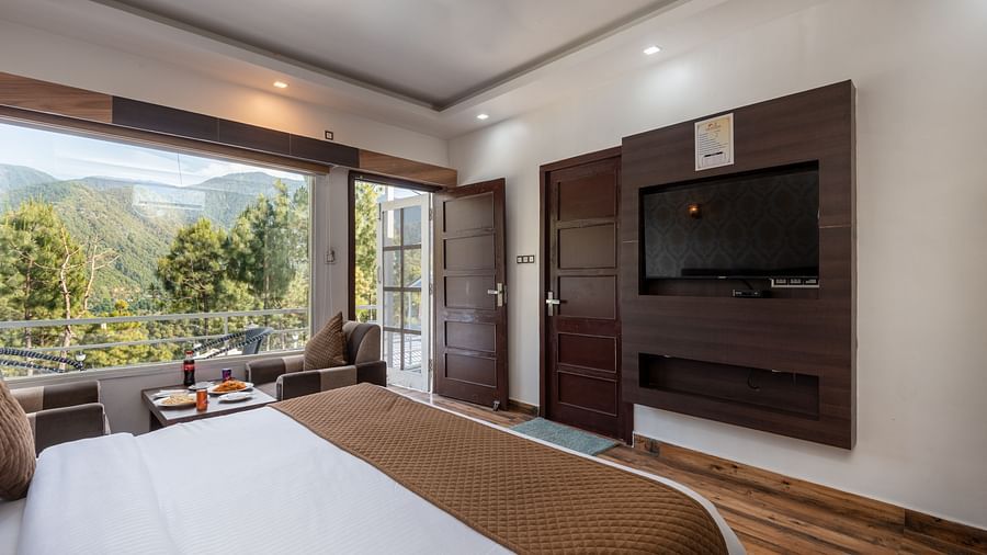  Cosy hotel room with a double bed, wall-mounted TV, wooden flooring, and an open door leading to a balcony with a scenic mountain and forest view - DLS Nature Trinket Resort, Dalhousie.