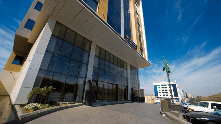 Corner view of Grand Iva Hotel's porch, driveway, and multi-storey structure's bottom few storeys, with a partially cloudy sky in the background,