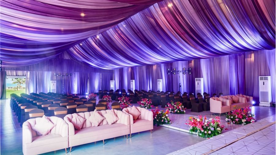 Decorated banquet hall with cream sofas and rows of chairs facing the stage featuring glowing purple decorated roof.