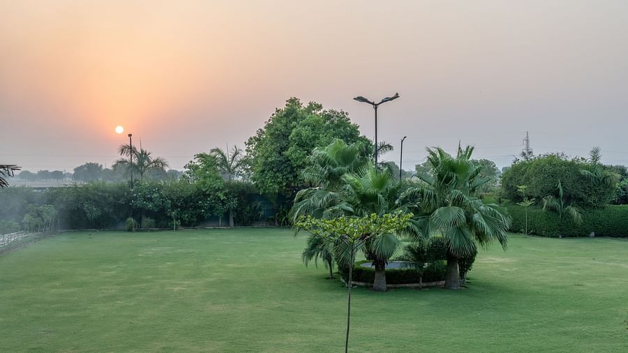 An extended view of the expansive open garden area in the premises of Hotel Highway King, Behror, seen with trees, grass, and glowing sun in the background.