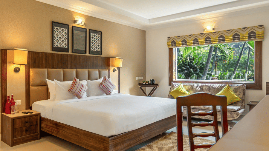 A hotel room with a bed, a desk, a sofa, and a large window overlooking a garden with palm tree at Mango Hill Resort, Coimbatore