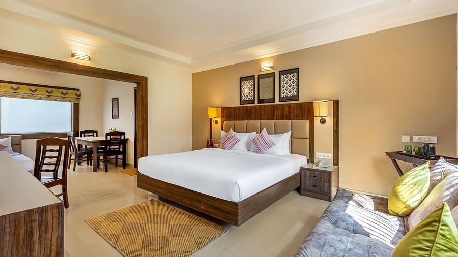 A hotel room with a queen bed, a sofa, and a doorway leading to a dining area with a table and chairs - Mango Hill Resort, Coimbatore