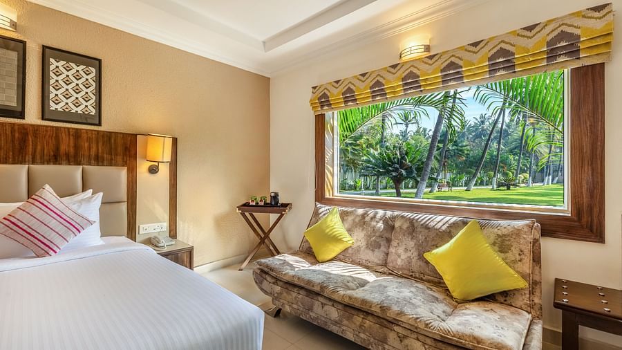 A hotel room with a queen bed, a patterned sofa, and a large window overlooking a garden with palm trees - Mango Hill Resort, Coimbatore
