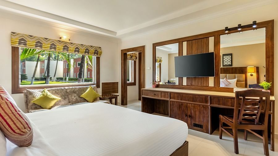 A bright bedroom with a bed with white linens, a wooden desk with a mounted TV, a patterned sofa with yellow pillows, and a window view of a tropical garden - Mango Hill Resort, Coimbatore